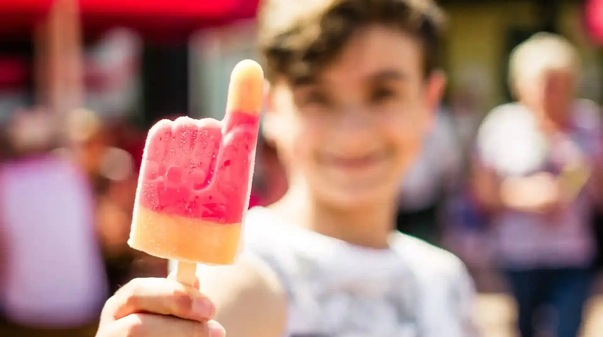 Auf dem Bild ist ein Junge zu sehen, der ein handförmiges Eis isst. Man sieht ihn im Hintergrund verschwommen grinsen. Entdecken sie erfrischen Angebote in der Rodelklause am Erlebnisberg in Altenberg.